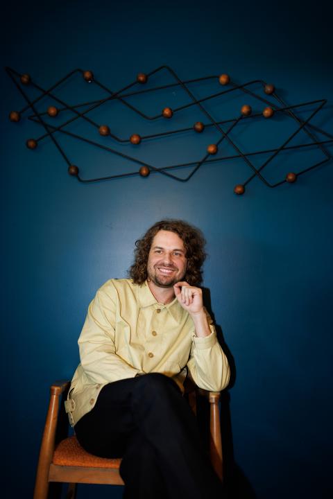 Kevin Morby sitting in a chair with a teal wall and an abstract linear sculpture above him
