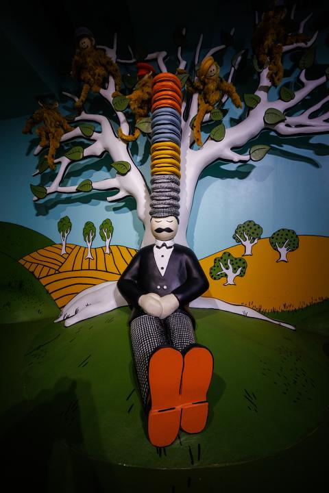 Whimsical sculpture of a man sitting against a tree wearing a large stack of hats