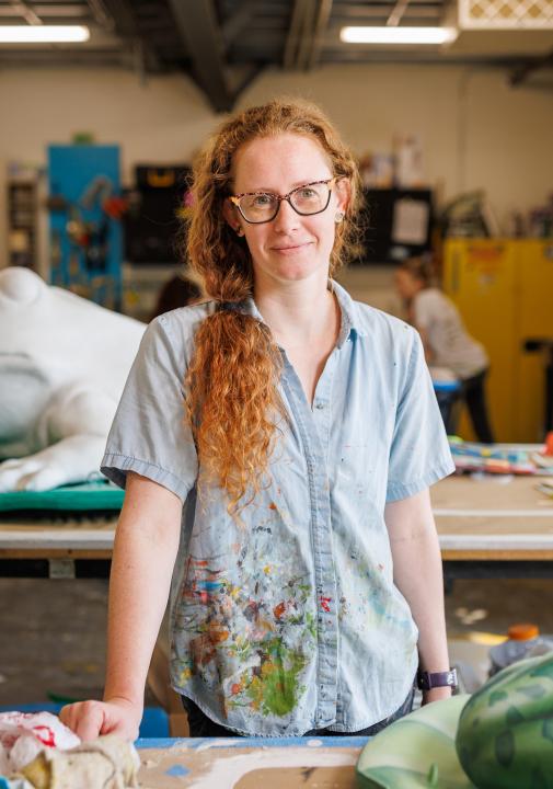 Artist Kelli Harrod standing in her workshop among in-progress artistic projects, including frog sculptures