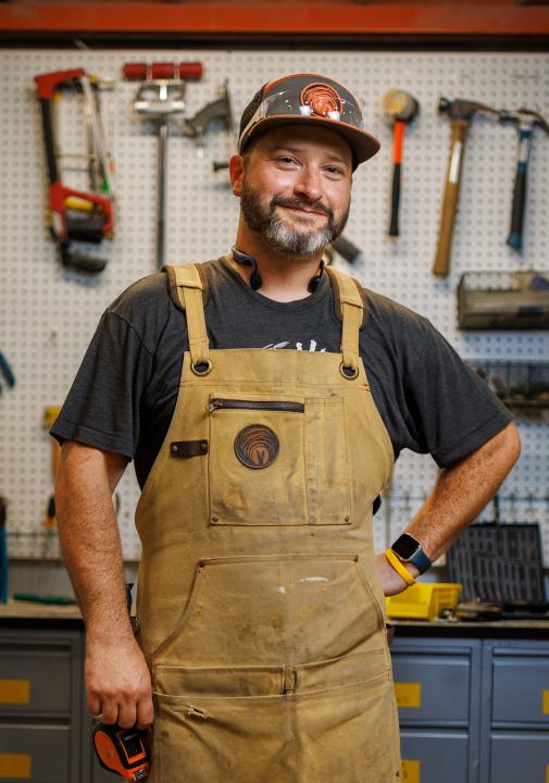 Artist Grand Keslo wearing a work apron, standing in a workshop with tools hanging up