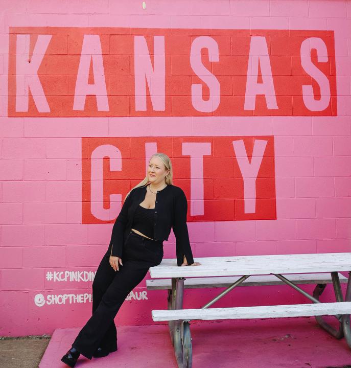 Sarah Sipple in an all-black outfit leaning against a white table, in front of a pink mural reading KANSAS CITY