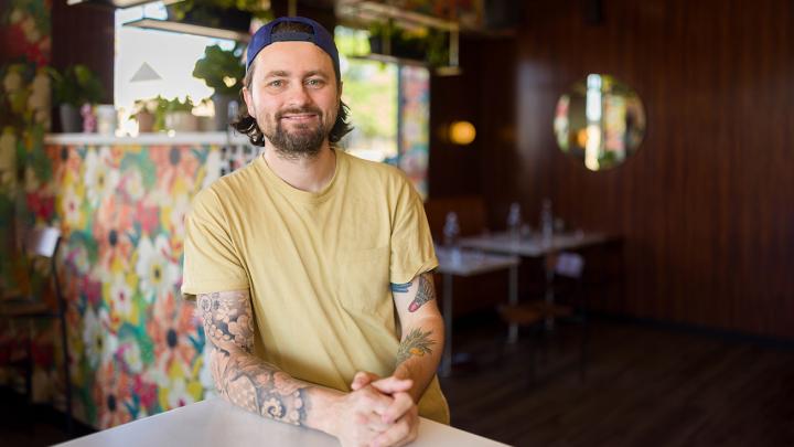 Jay Sanders wearing a yellow shirt and baseball cap, standing at a bar.