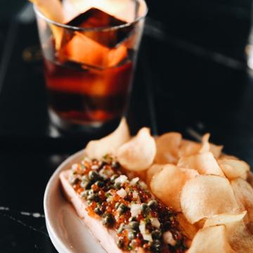 A dish of salmon topped with a spread and chips, alongside an old fashioned cocktail.