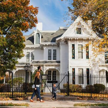 Parents and young child walking in front of a large white house surrounded by autumn trees