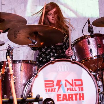 Woman playing a drumset with a logo saying Band That Fell to Earth